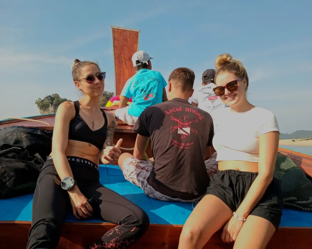 5 people in the bow area of a traditional longtail boat. The 2 women closest are facing the camera and smiling. One giving a thumbs up , The three men behind them area ll facing away, all are wearing Local Diving t shirts