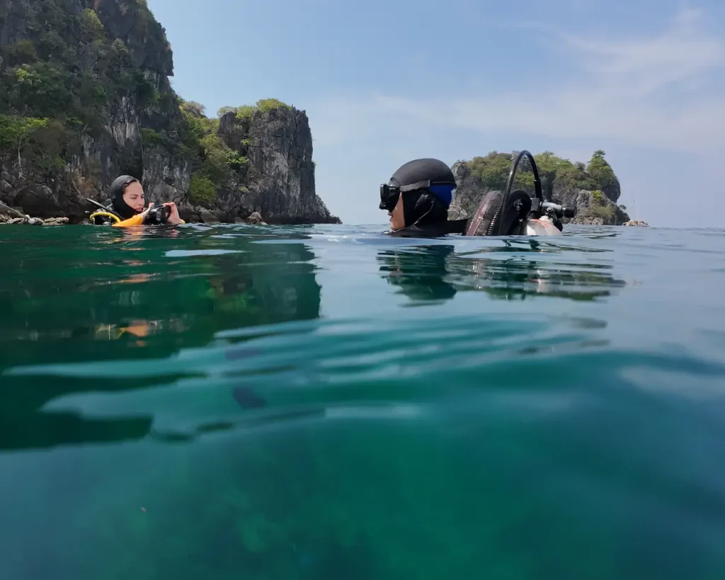 two scuba divers on the surface of calm water preparing to dive. One is checking her mask. With small rocky islands in the background