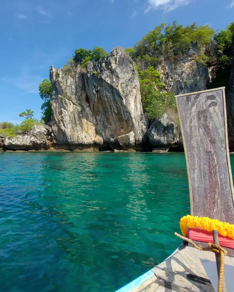 the high prow of a traditional longtail boat from Krabi approaching a rock island there is some vegetation growing on the rocks. the sea water looks calm and clear, the sky is blue