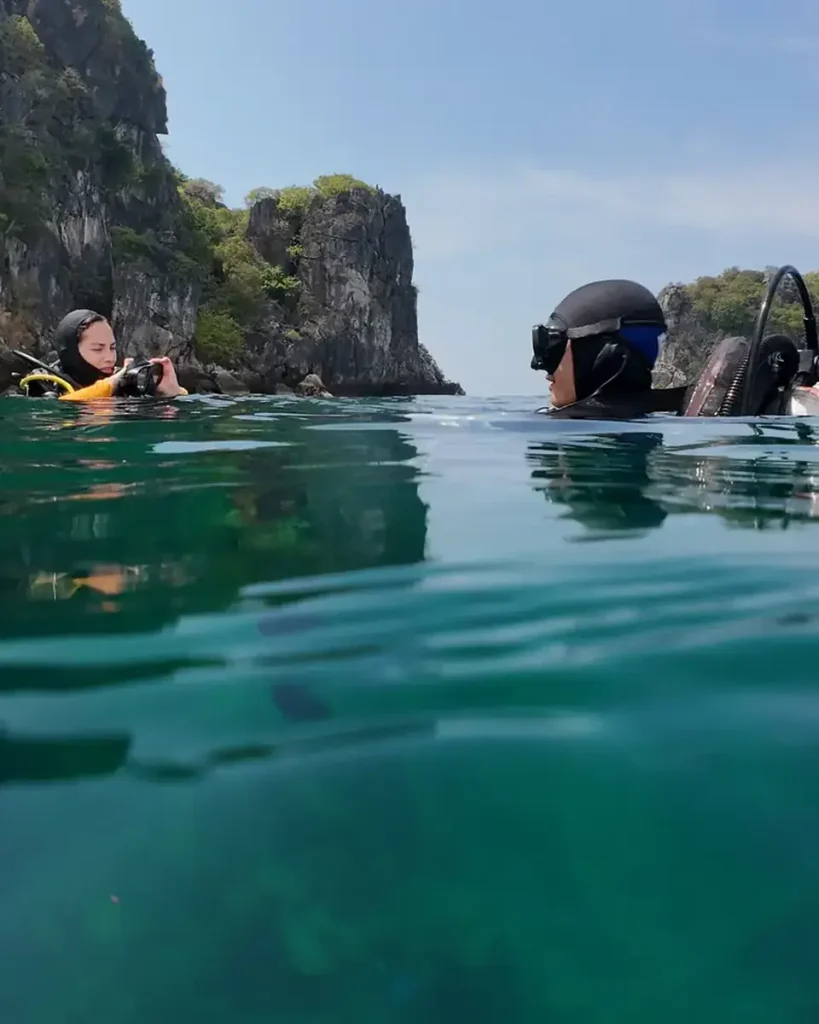 two scuba divers on the surface of calm water preparing to dive. One is checking her mask. With small rocky islands in the background