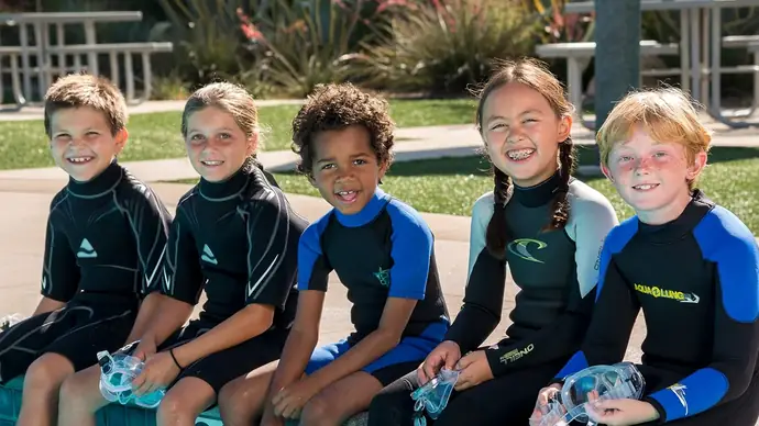 a group of 5 children sit next to each other on a bench. they are in wetsuits and holding diving masks. Getting ready for a PADI bubblemaker junior scuba course