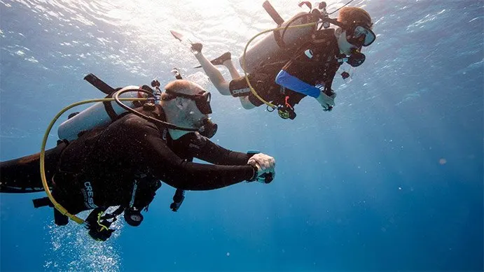 two divers swim by, in the foreground a man swims with his hands clasped in front of him, he's looking up slightly to a younger man who is porbably his SCUBA diving student.