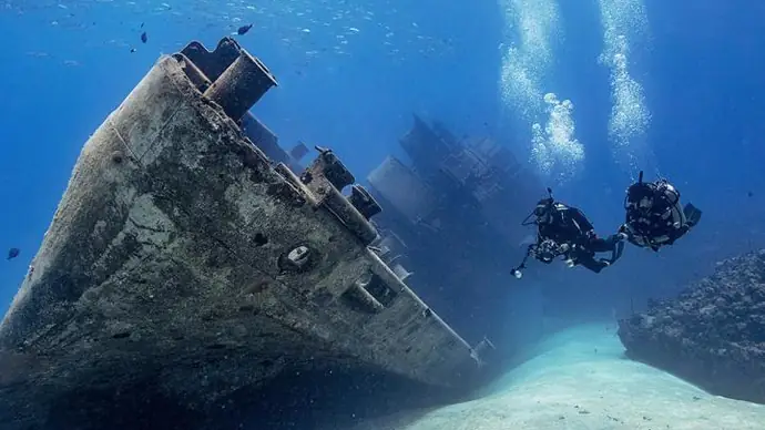 2 scuba divers underwater with a shipwreck. one diver has a large camera housing with two strobe arms, the water is clear. the wreck is large and mainly intact. the divers are near the bow