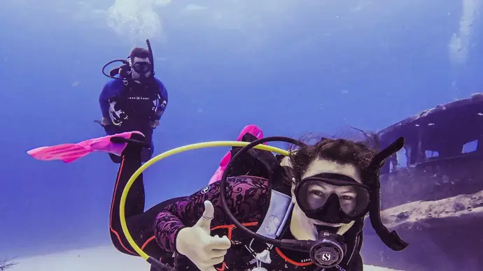 a female scuba diver in the foreground looks ahead and signals thumbs up. behind her a male diver is swimming