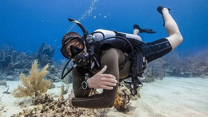 a person in a scuba gear underwater, diving over a sead bed of fine sand and outcrops of coral.. the diver's arms are folded in front of him as he looks at the camera and smiles, although hard to see behind the mask and regulator