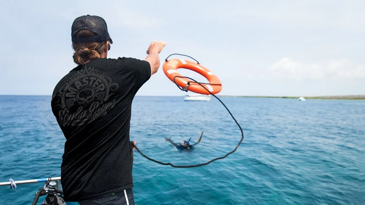 a man on a boat, viewed from behind has thrown a bright orange circular life preserver attached to a rope to a man in the water who is reaching out to catch it