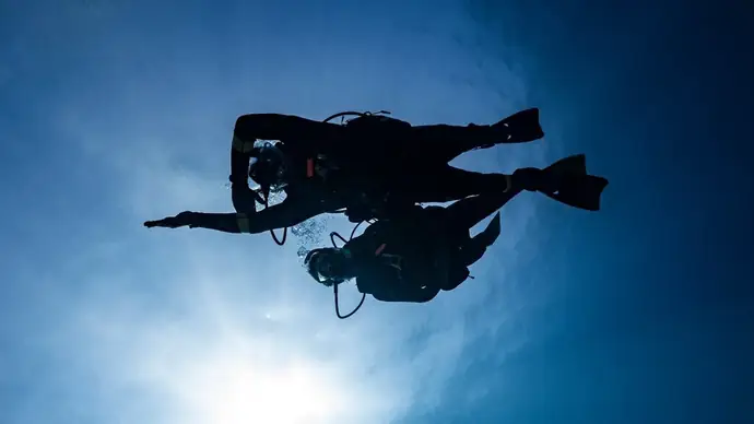 viewed from below with the water surface and sun above. there are 2 scuba divers silhouetted in the water. one with their right arm stretched out ahead of them they swim horizontally. the other diver is close by supervising, instructing an exercise on the underwater navigation specialty course.